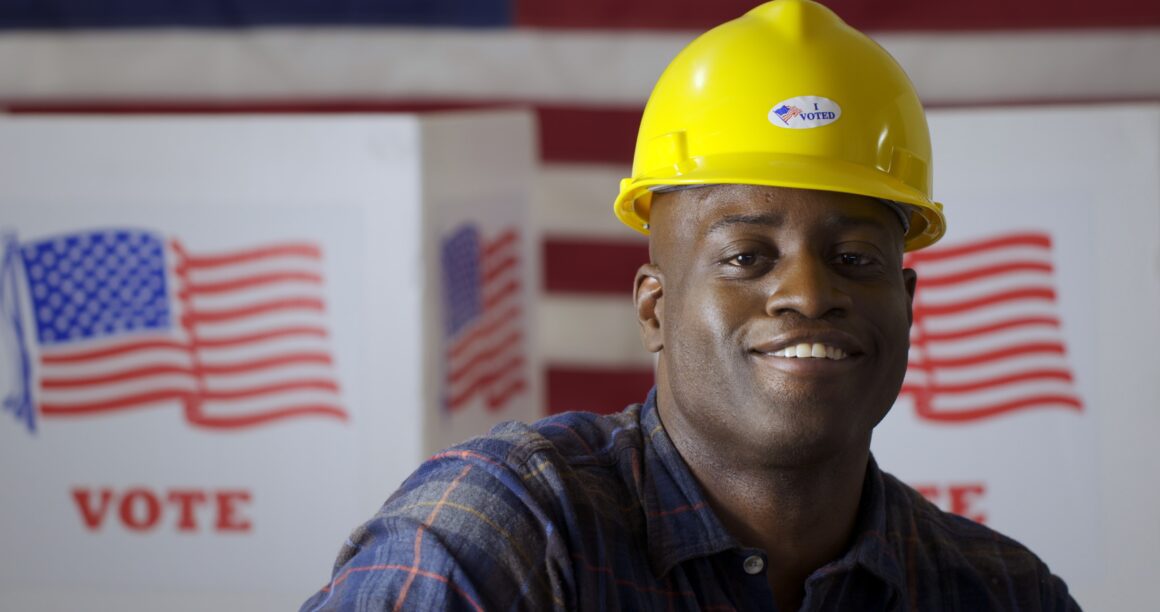Close up African American man in plaid shirt wearing hard hat wi Smiling construction worker, I Voted sticker.